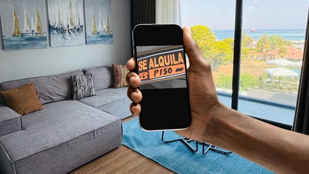 Mano sosteniendo un móvil con un cartel de “se alquila piso” dentro de un apartamento con vistas al mar