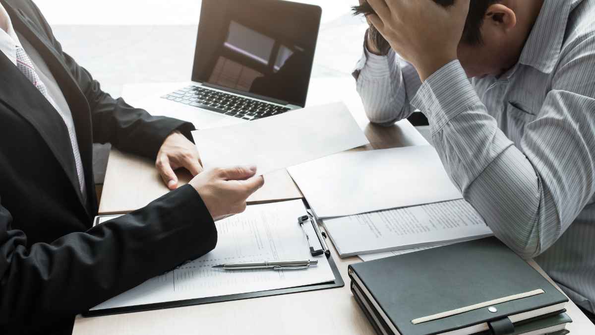 Empleado recibiendo notificación laboral mientras otro trabajador muestra signos de preocupación en una reunión