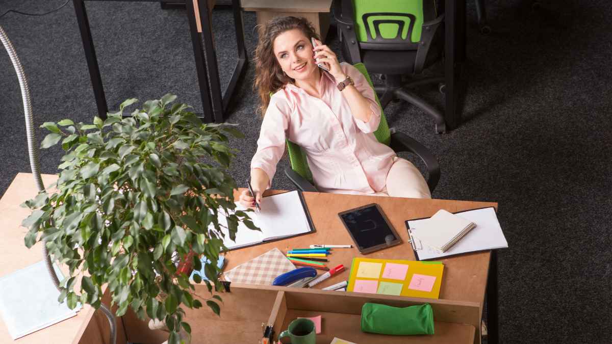 Mujer trabajando en una oficina mientras habla por teléfono y toma notas en su escritorio.