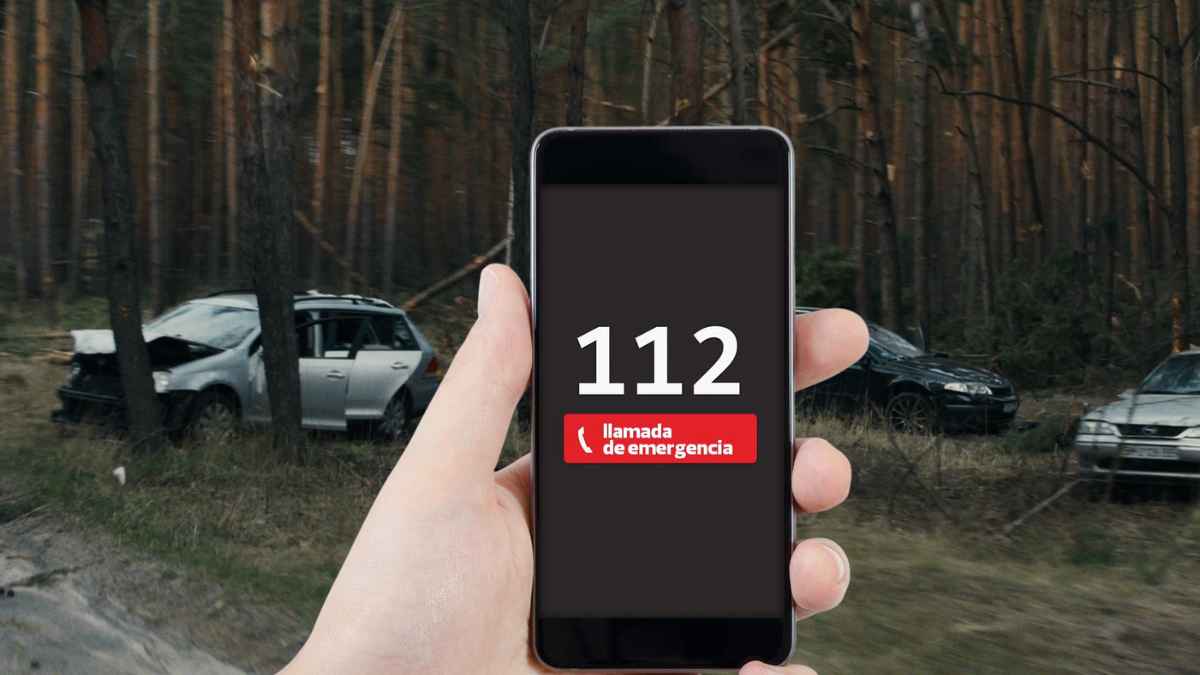 Teléfono móvil mostrando una llamada al 112 con un accidente de tráfico al fondo en una carretera forestal