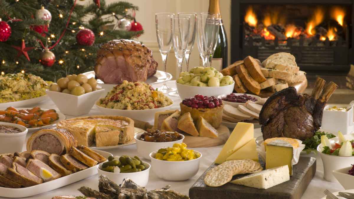 Mesa de cena navideña con alimentos típicos como carnes, quesos, entrantes y postres junto a chimenea encendida y árbol de Navidad