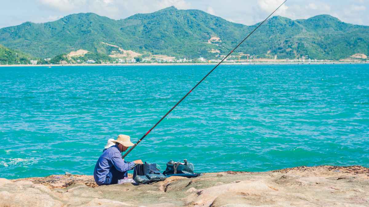 Persona pescando en la orilla del mar con montañas al fondo en un día soleado