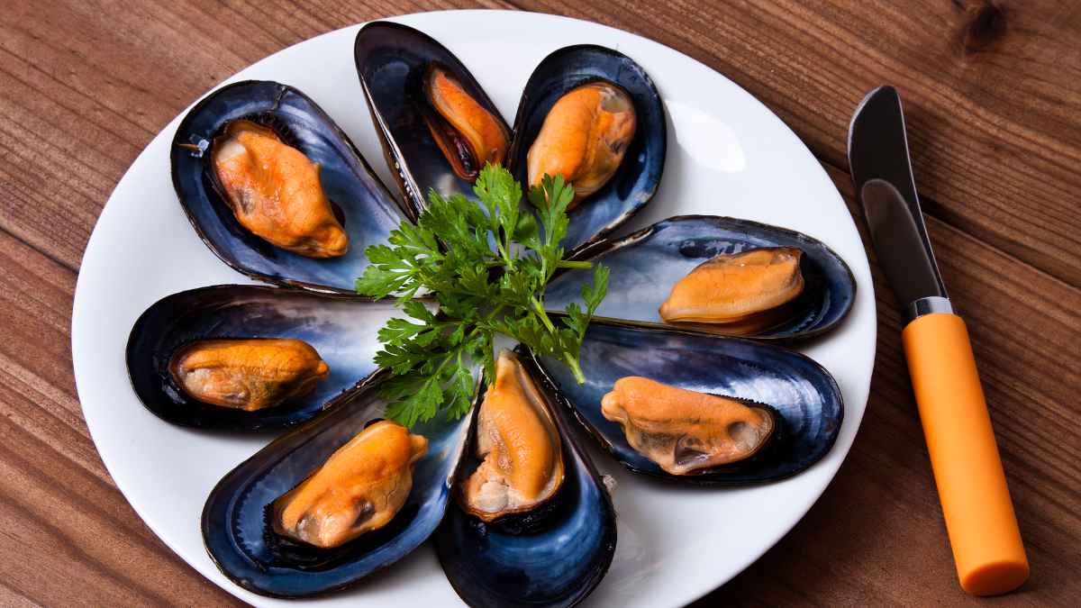 Plato de mejillones cocidos abiertos con perejil, servidos en un plato blanco sobre mesa de madera, con cubiertos al lado.
