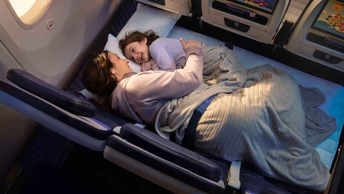Madre y niña descansan tumbadas en una fila de asientos de avión convertida en cama en clase económica, con manta y almohada.