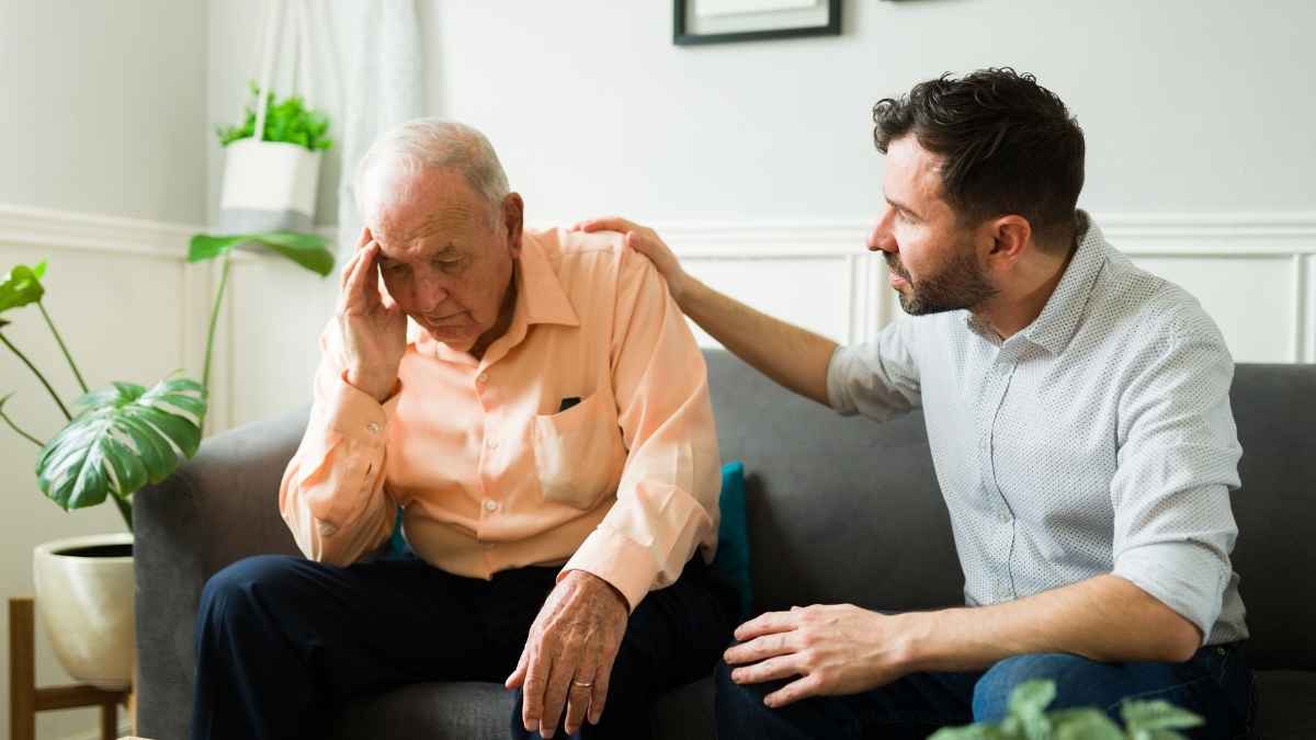 Hombre mayor sentado en un sofá con gesto de malestar mientras otro hombre le apoya con la mano en el hombro, imagen de baja laboral por ictus