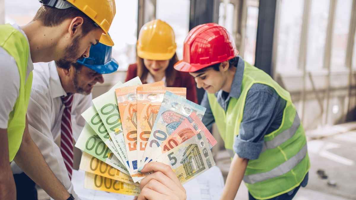 Grupo de trabajadores con casco de seguridad mostrando billetes de euro en una construcción, representando la relación entre el trabajo y las ayudas económicas del SEPE