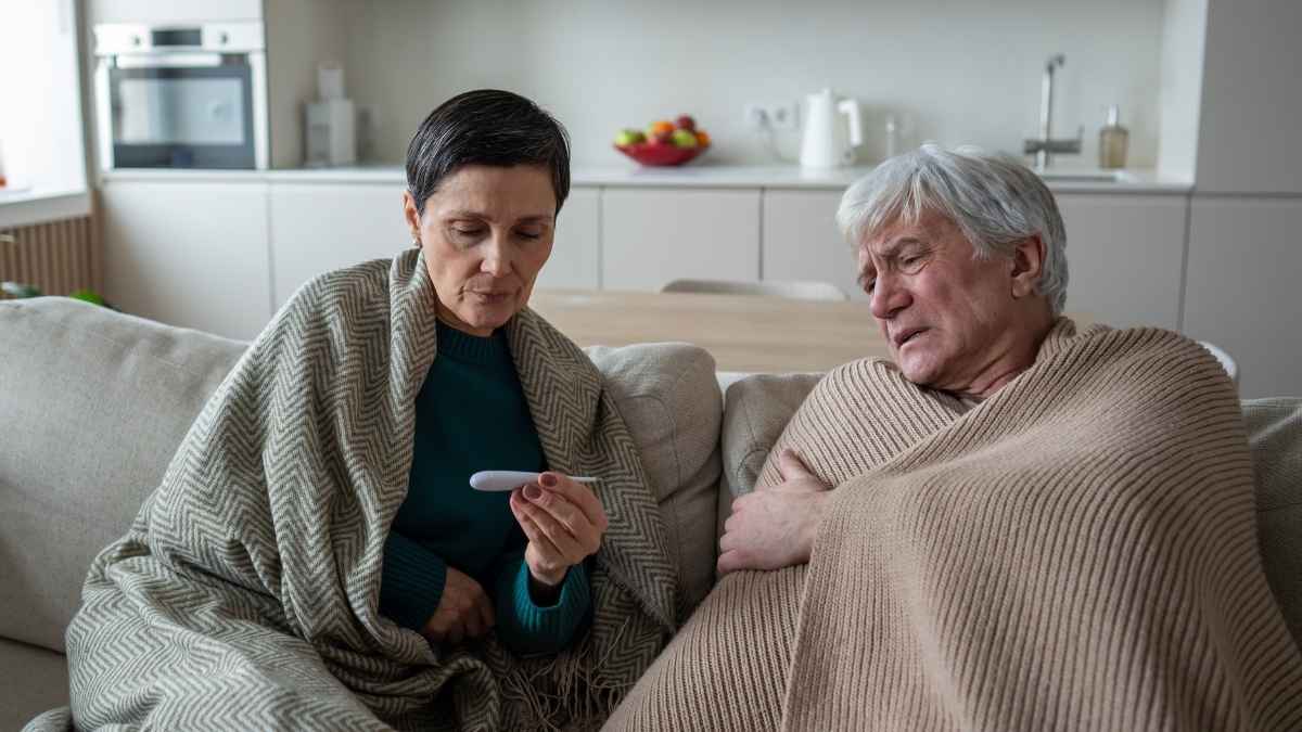 Pareja de adultos mayores cubiertos con mantas, revisando el termómetro en casa mientras descansan en el sofá