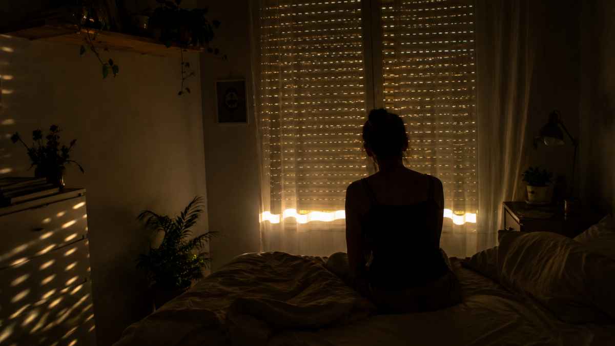 Persona sentada en la cama con la persiana bajada y rayos de sol entrando por los huecos