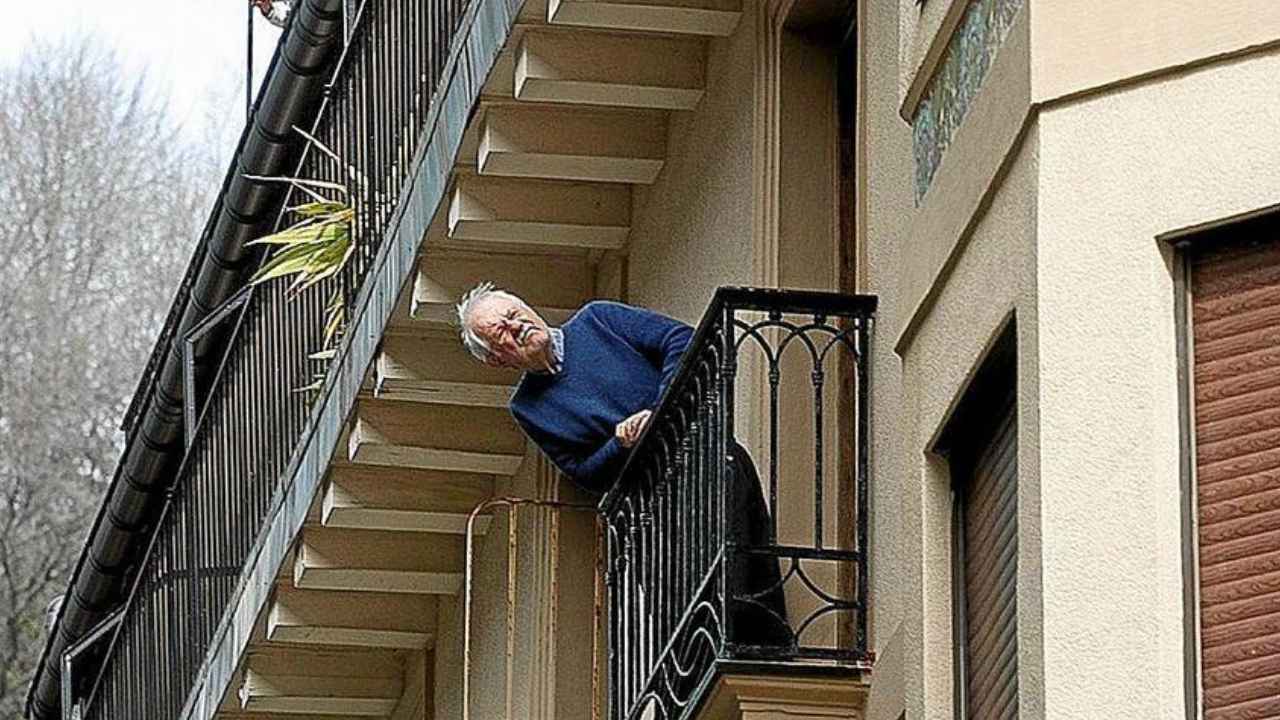 Hombre mayor asomado a un balcón de un edificio residencial.