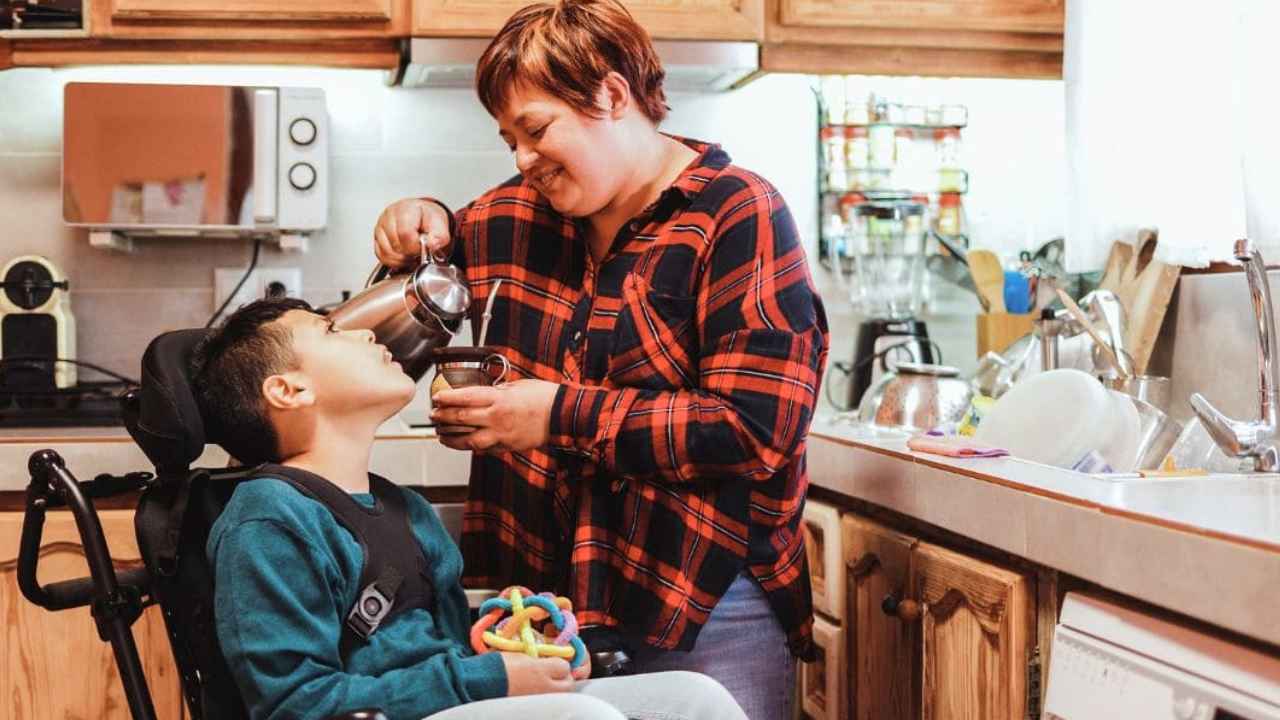 Una mujer ayuda a un niño en silla de ruedas en una cocina, imagen de los cuidados familiares a personas dependientes.