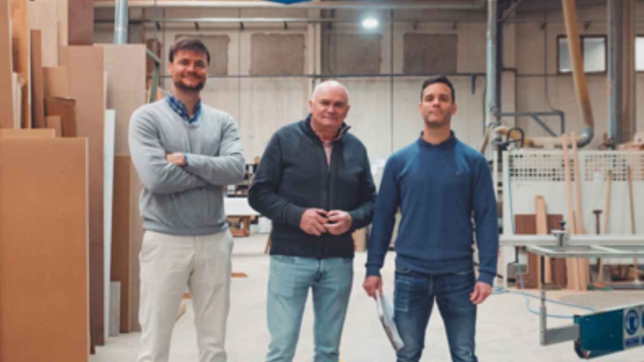 Tres hombres posan en un taller de carpintería junto a tableros de madera y maquinaria.