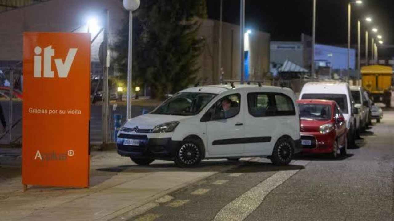 Varios vehículos estacionados junto a una estación de ITV durante la noche, con un cartel naranja de inspección técnica.