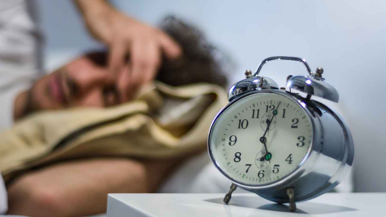 ALT: Despertador negro en primer plano junto a una mujer dormida en la cama. Despertador negro en primer plano junto a una mujer dormida en la cama.