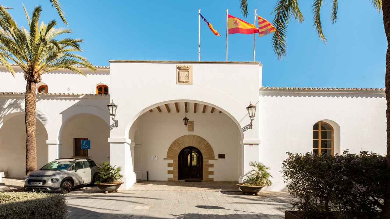 Fachada blanca de un edificio institucional con arcos, banderas y palmeras junto a la entrada.