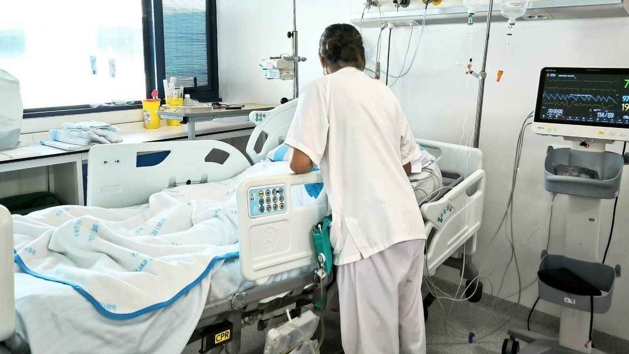 Profesional sanitaria atendiendo a un paciente hospitalizado en una habitación con cama clínica y monitor de constantes vitales.