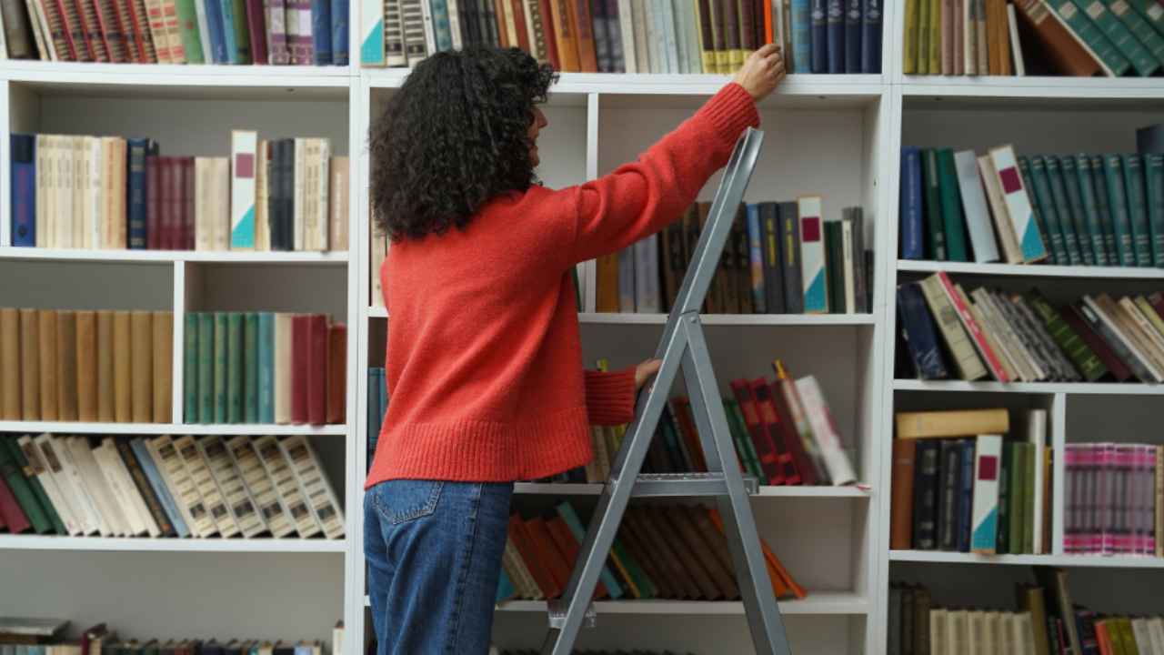 Mujer colocando libros en una estantería de biblioteca con ayuda de una escalera.