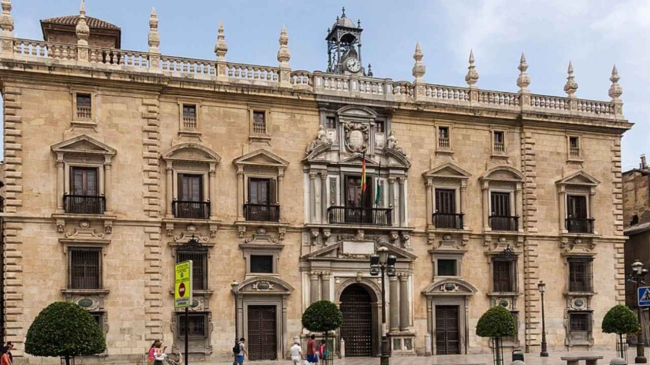 Fachada de la Real Chancillería de Granada con balcones, banderas, árboles y peatones ante el edificio.